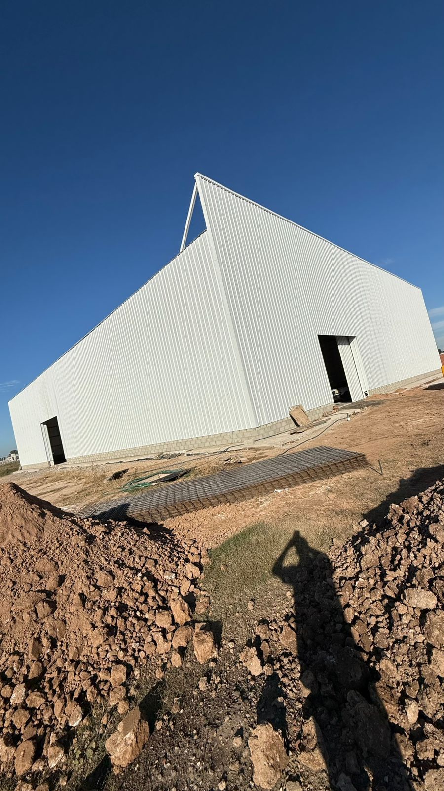 Construcción de un gran edificio metálico blanco bajo un cielo azul despejado. El edificio tiene un tejado a dos aguas y dos puertas abiertas.