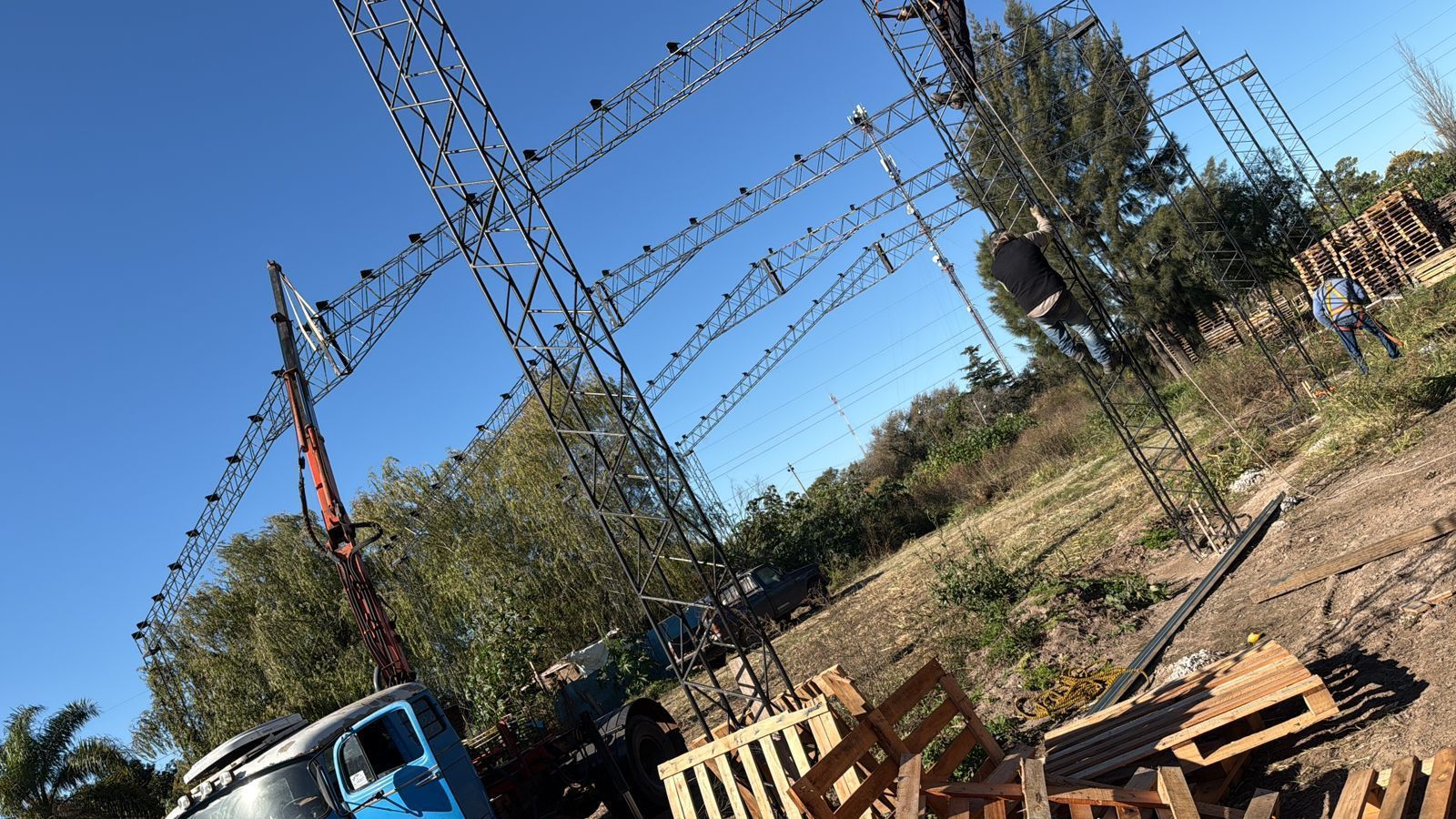 Una grúa cerca de una gran estructura metálica bajo un cielo azul. Hay palés de madera dispersos en primer plano.