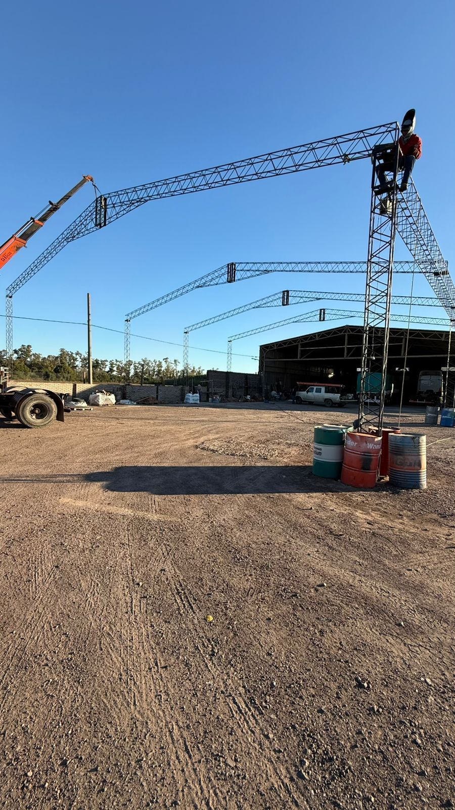 Obra con grandes grúas bajo un cielo azul despejado. El entorno es un terreno de grava con estructuras industriales.