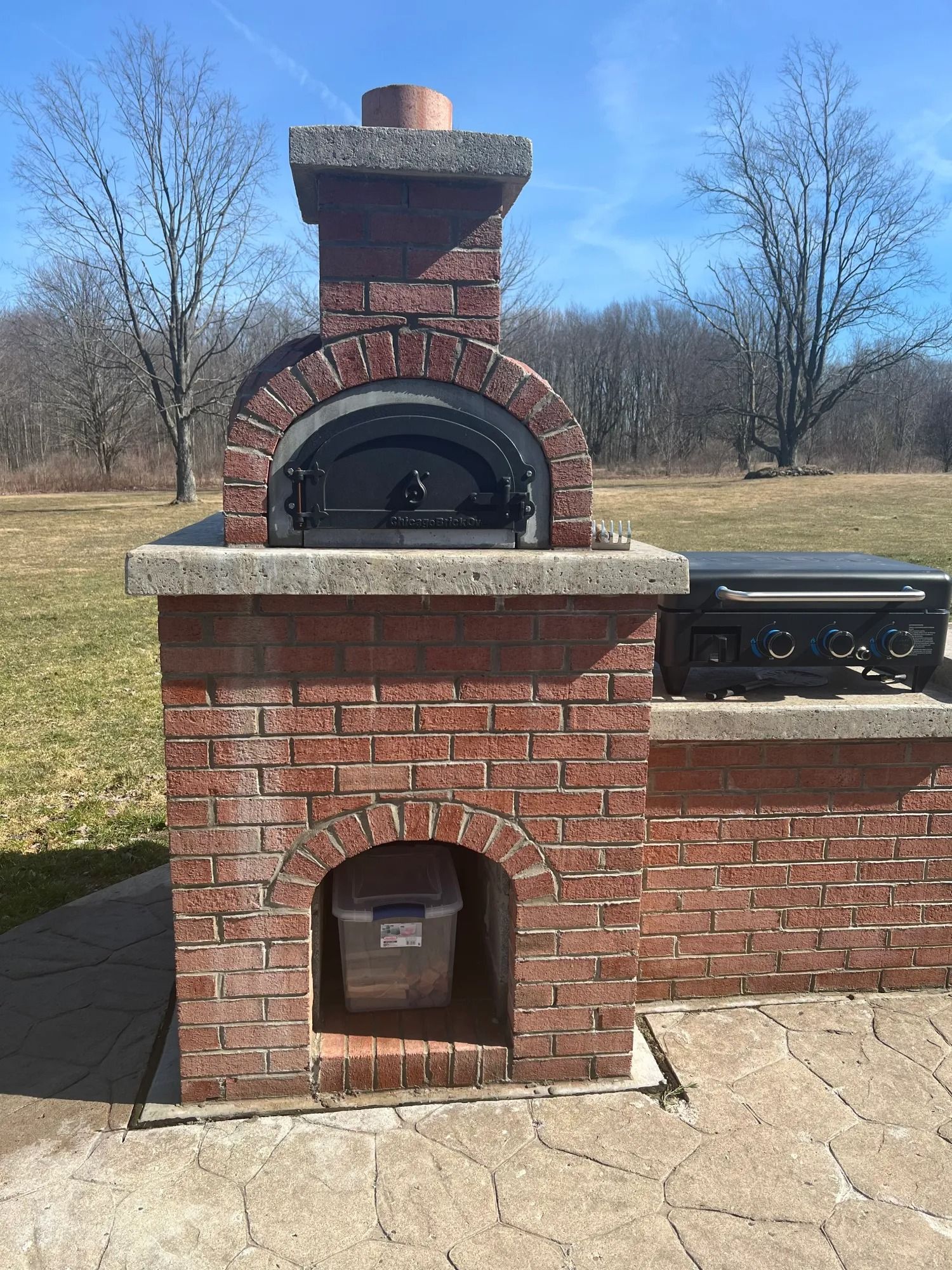 Brick outdoor oven with chimney and adjacent grill on a patio.