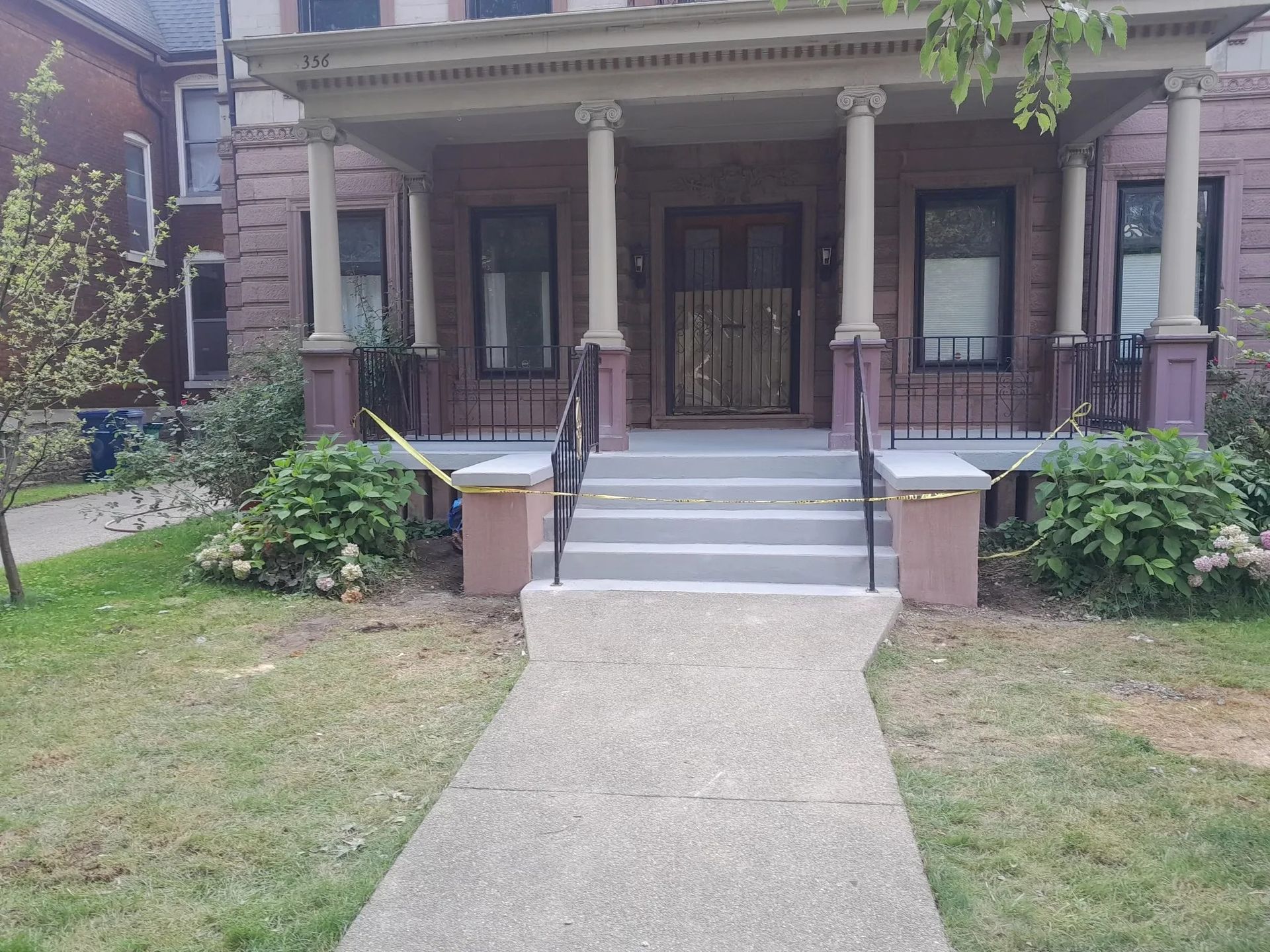 A gray concrete walkway leads to a house with a front porch and steps, with bushes on either side.