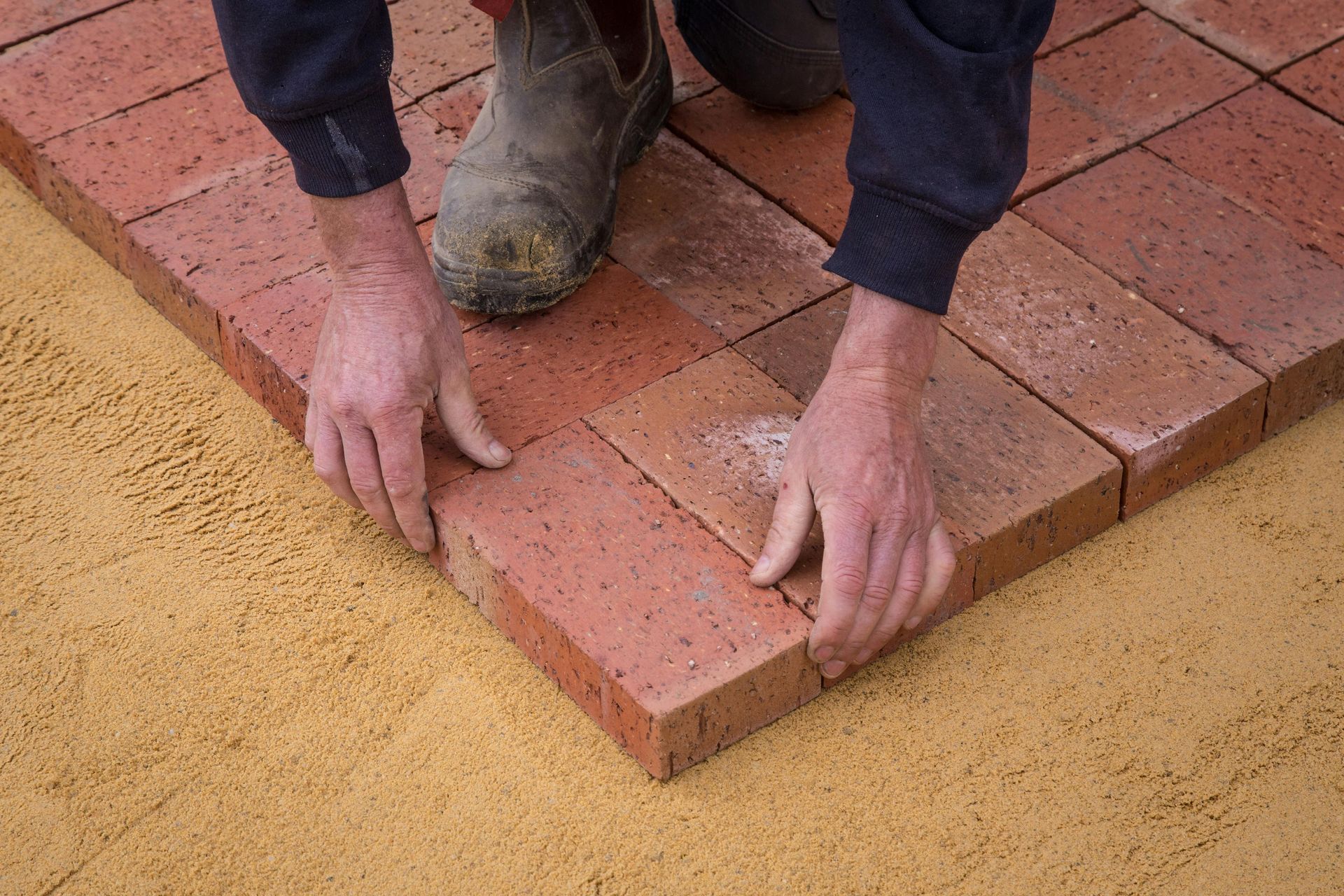 Person laying red bricks on a sand base. Hands placing brick; work boots visible. Outdoor setting.