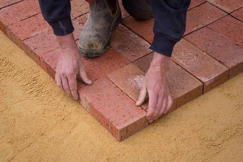 Person laying red bricks on a sand base. Hands placing brick; work boots visible. Outdoor setting.