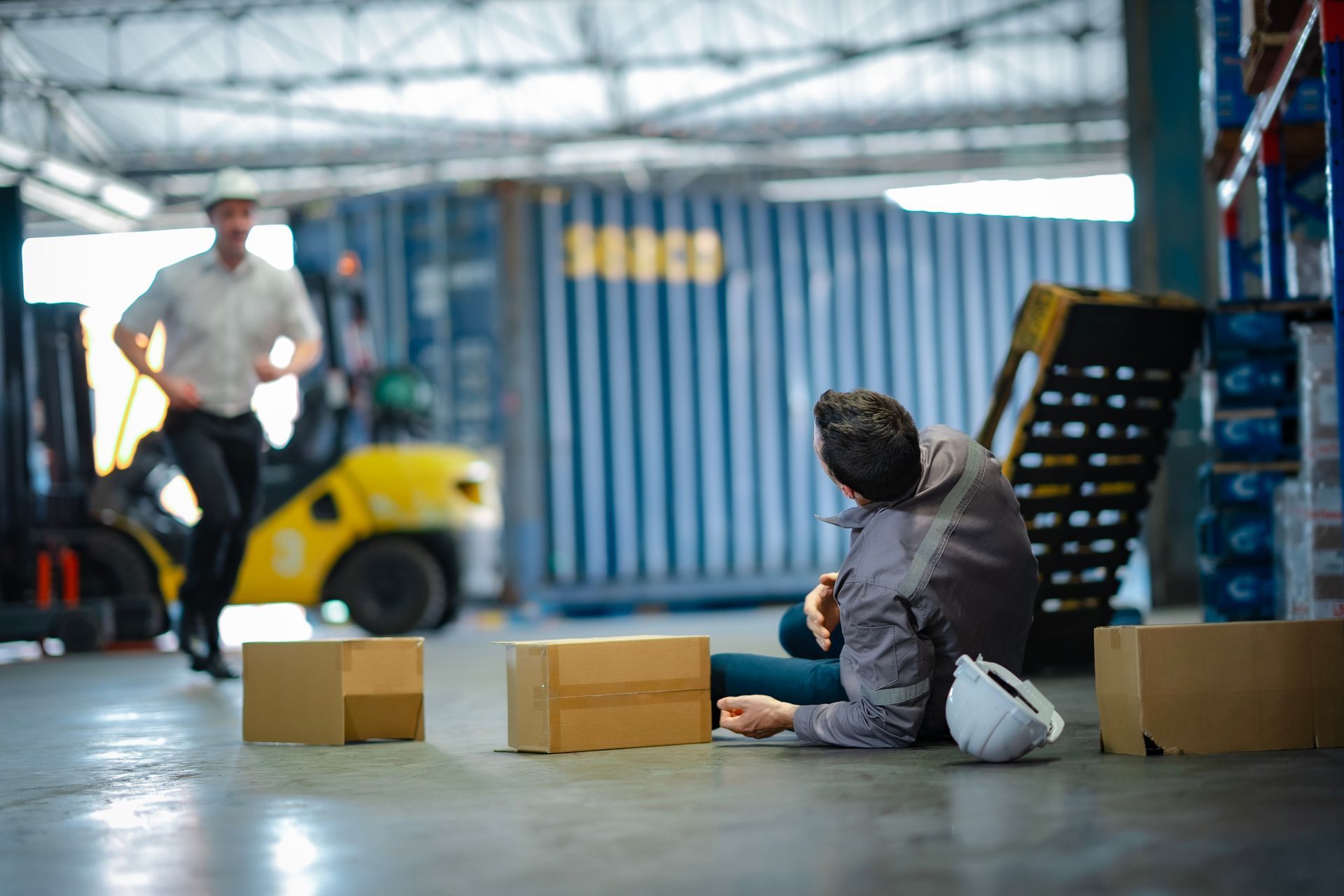 A male worker lies on the ground next to some boxes and a ladder after falling, inside a warehouse.