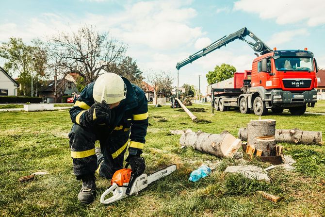 Firefighter in gear kneeling with a chainsaw near logs and a red crane truck.