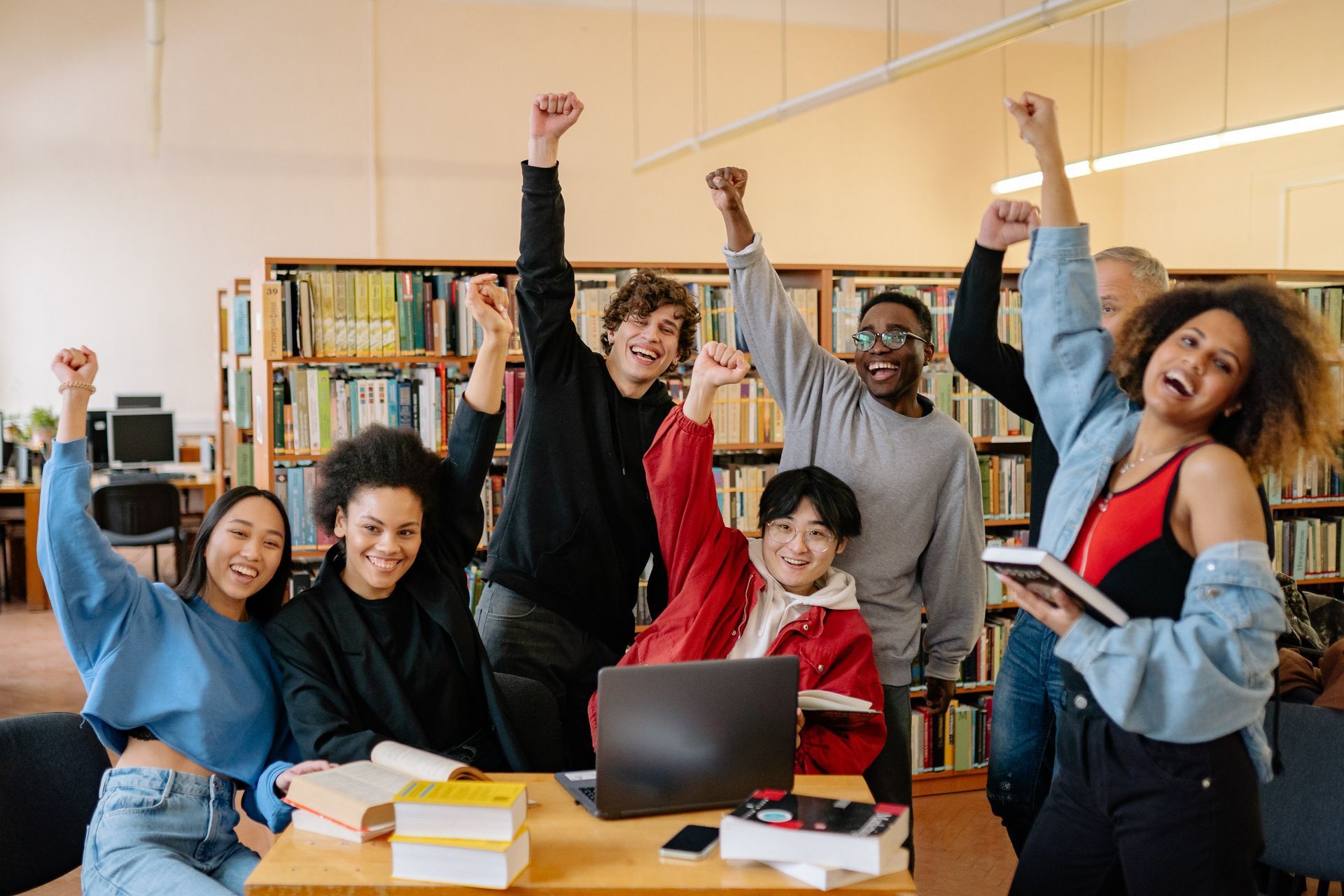 A group of young people are sitting at a table in a library with their arms in the air.