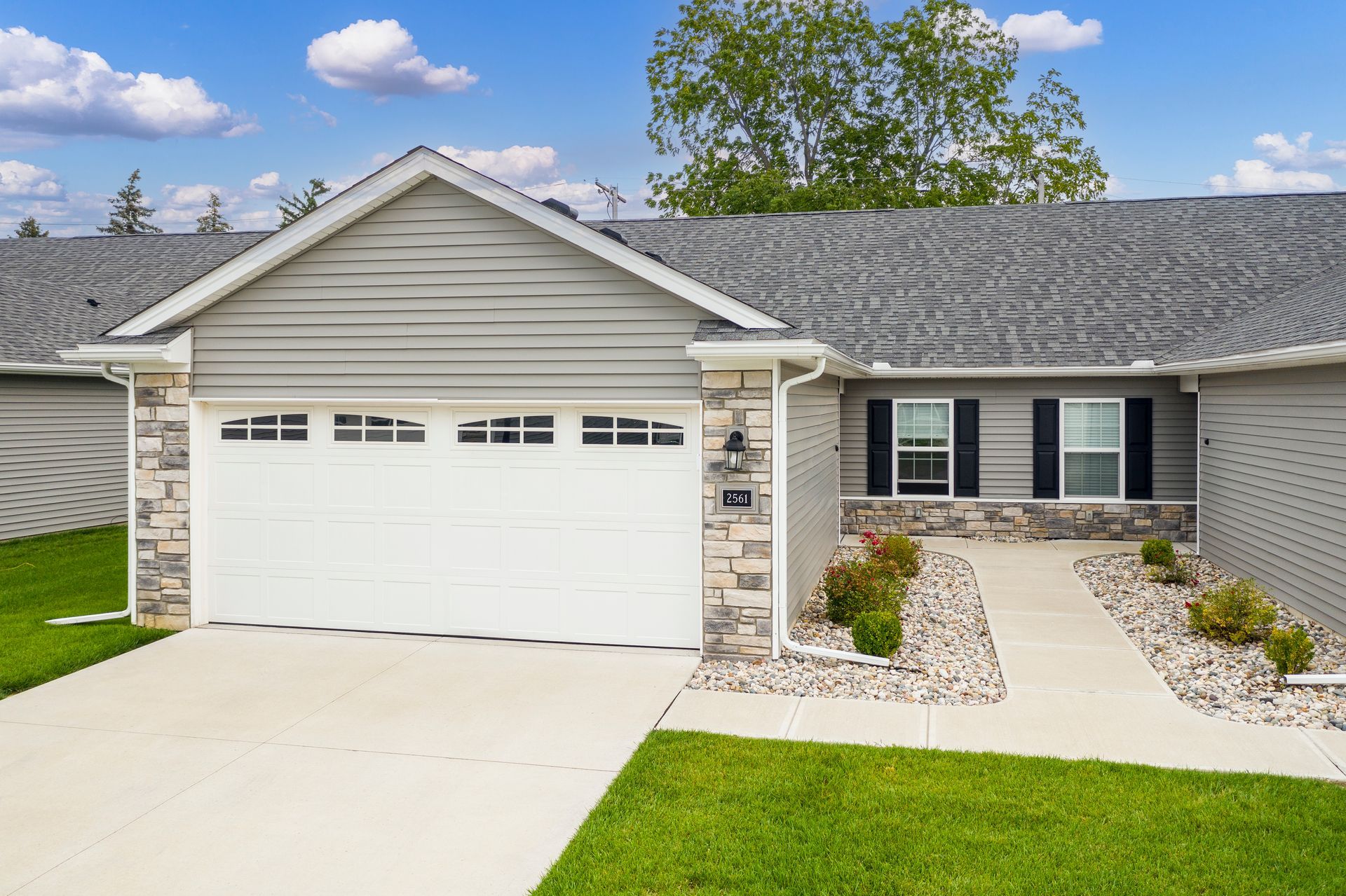Gray house with a white garage door, stone accents, and a walkway. Green lawn and blue sky.