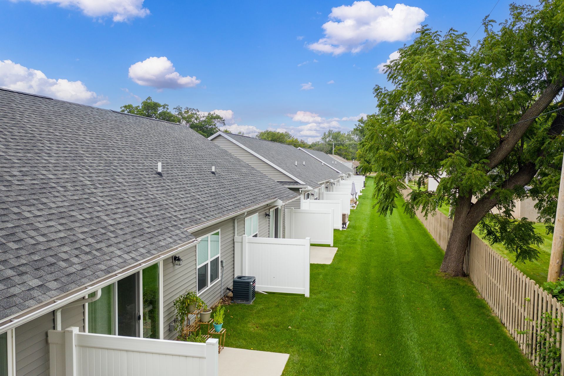 Row of townhomes with gray roofs and white fences, green lawn, tree, and blue sky.