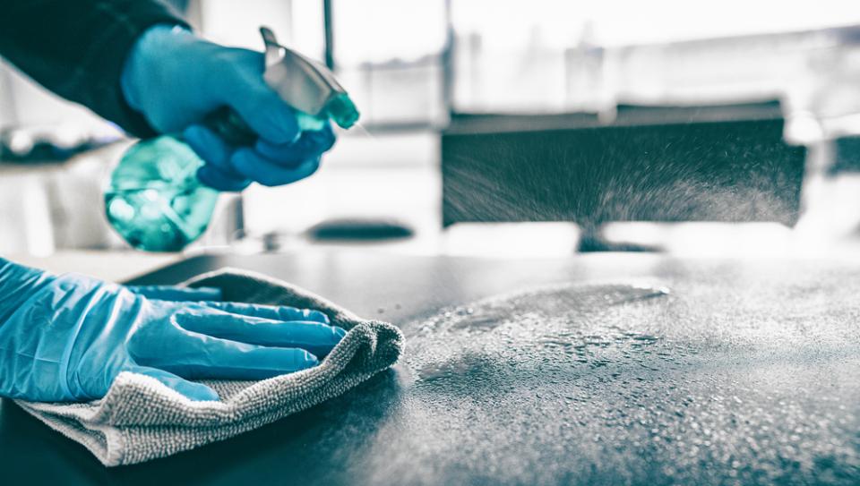 Una persona che indossa guanti blu sta pulendo un tavolo con un panno e uno spruzzatore.