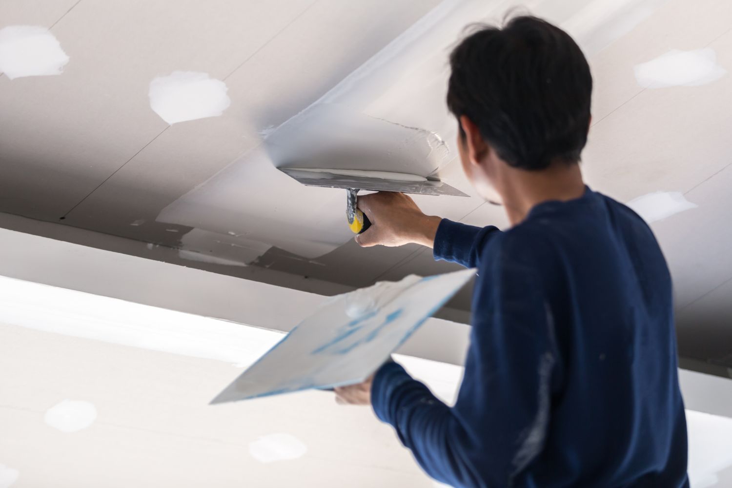 A man is drywalling on a ceiling with a tool.