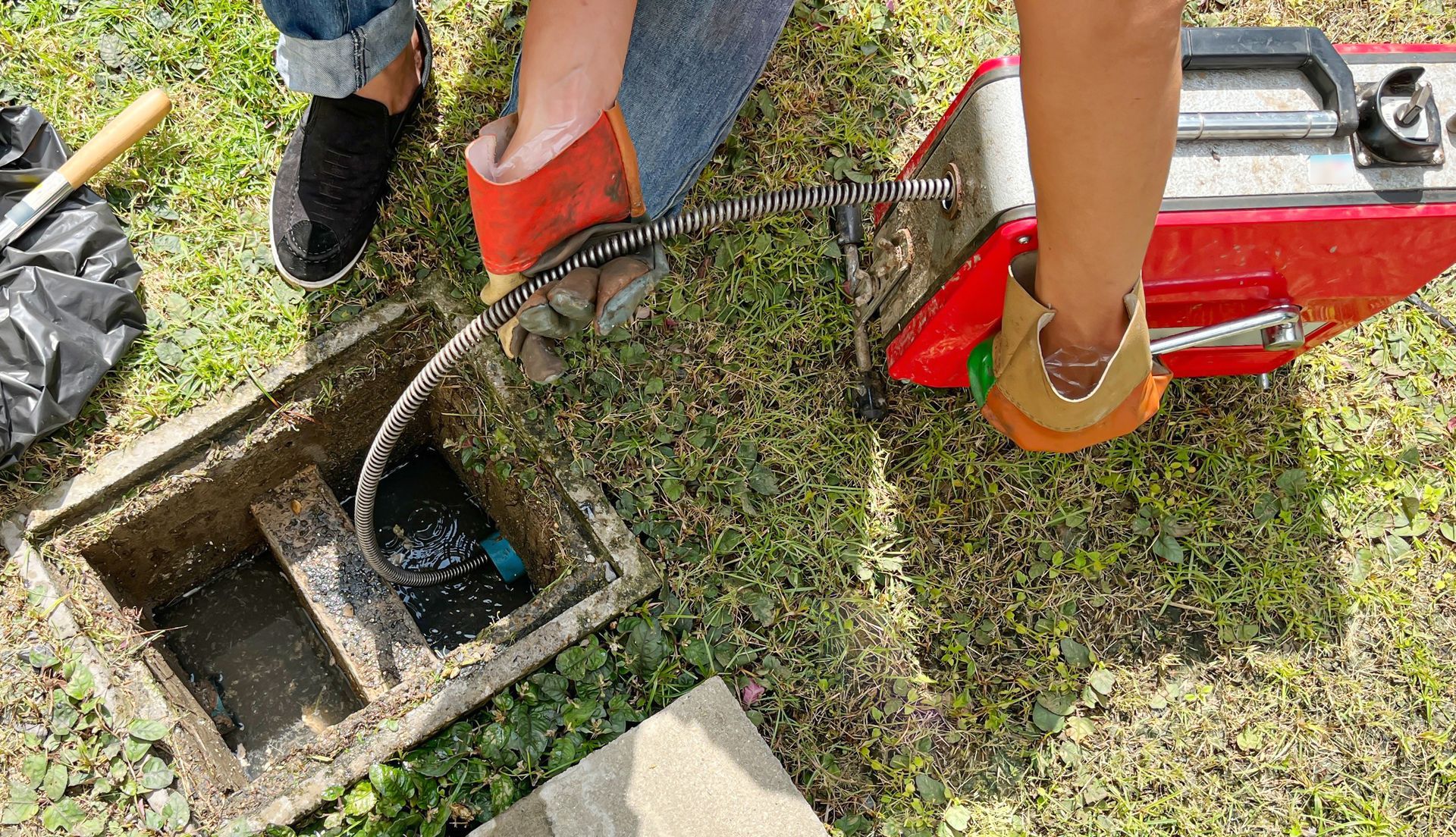 Someone using a drain snake machine to clear a clogged outdoor drain on grassy ground with tools