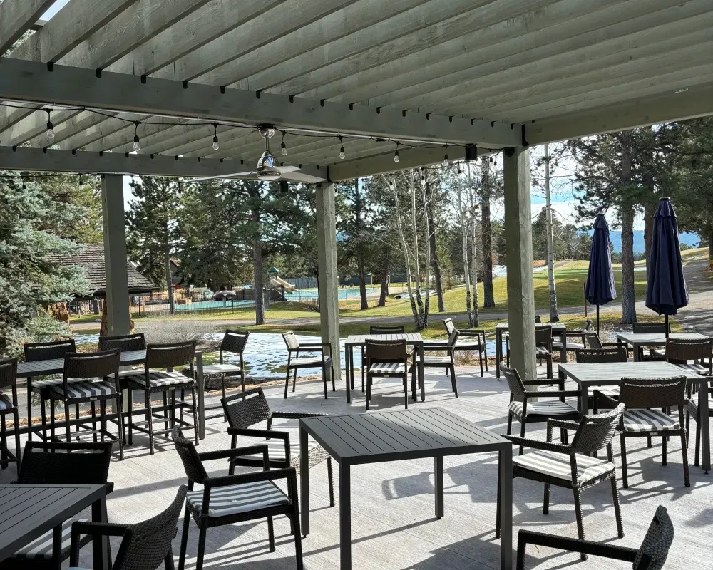 Outdoor patio with tables and chairs, under a pergola. View of trees and a golf course in the distance.