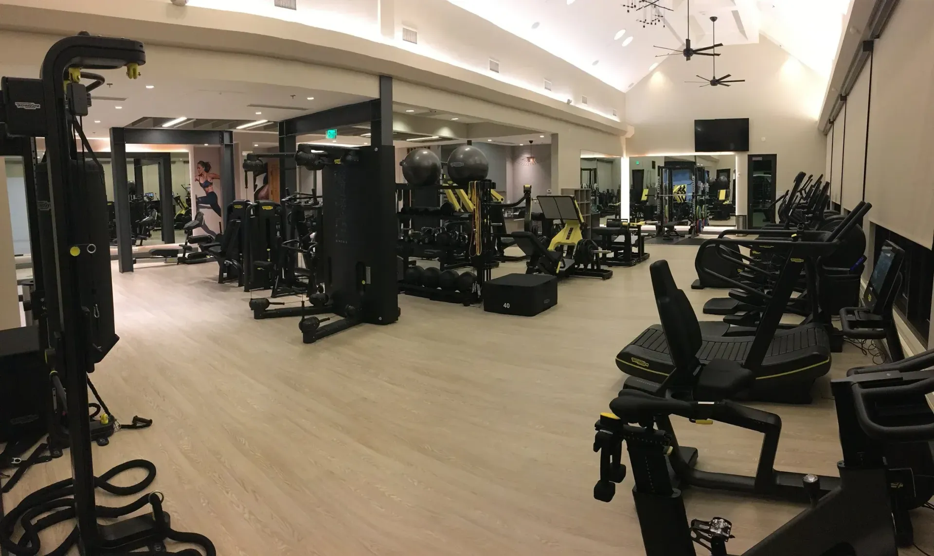 Gym interior with exercise equipment on light-colored floor; mirrors, and high ceiling.