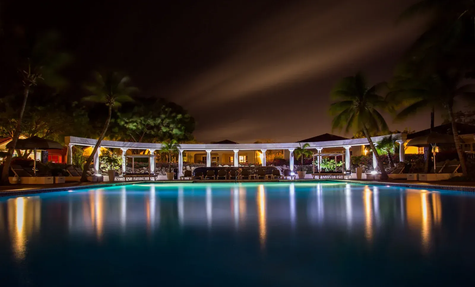 A large swimming pool is lit up at night with palm trees in the background.
