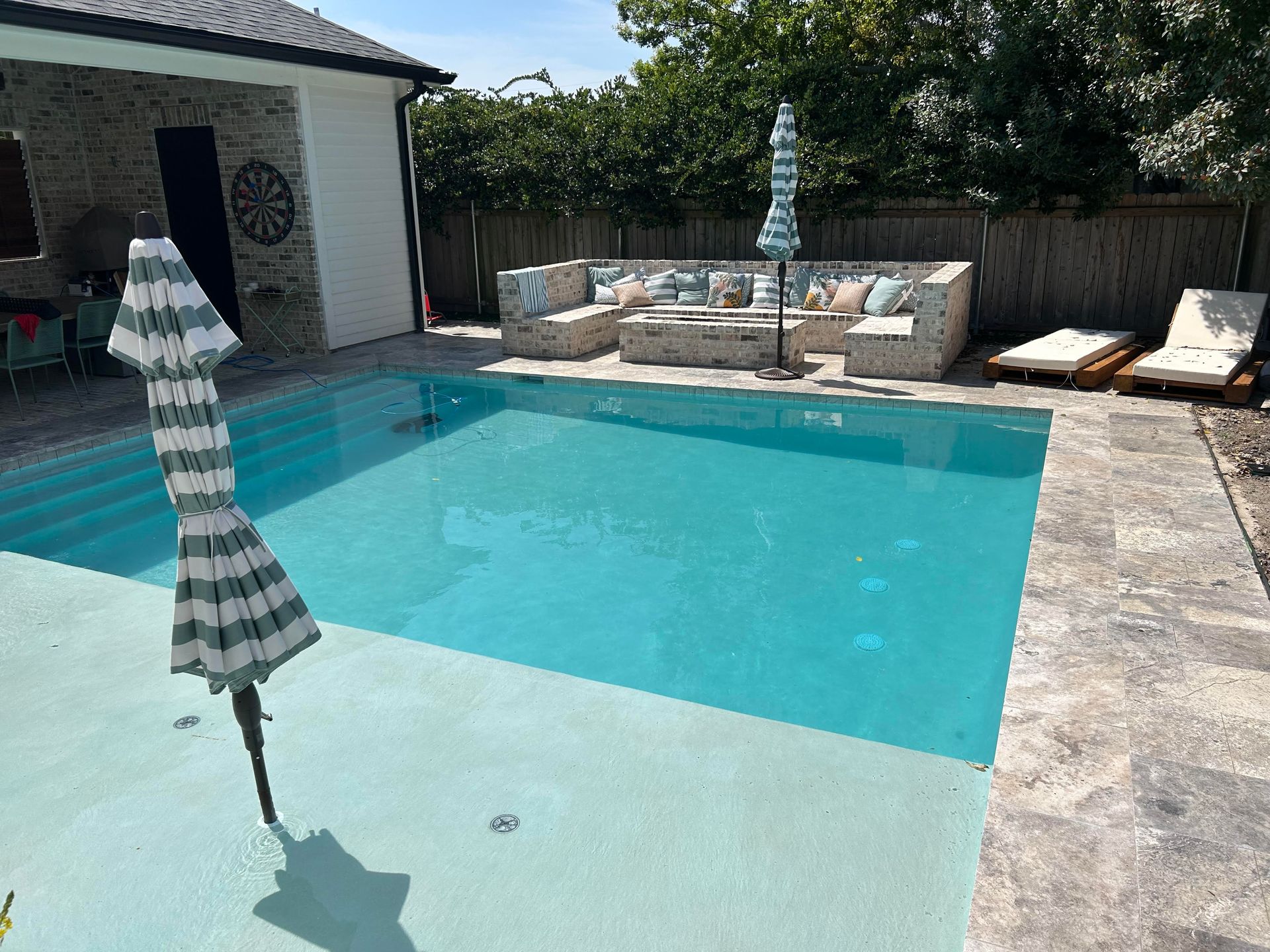 A backyard patio featuring a square pool, a stone fire pit area with seating, and lounge chairs on a sunny day.
