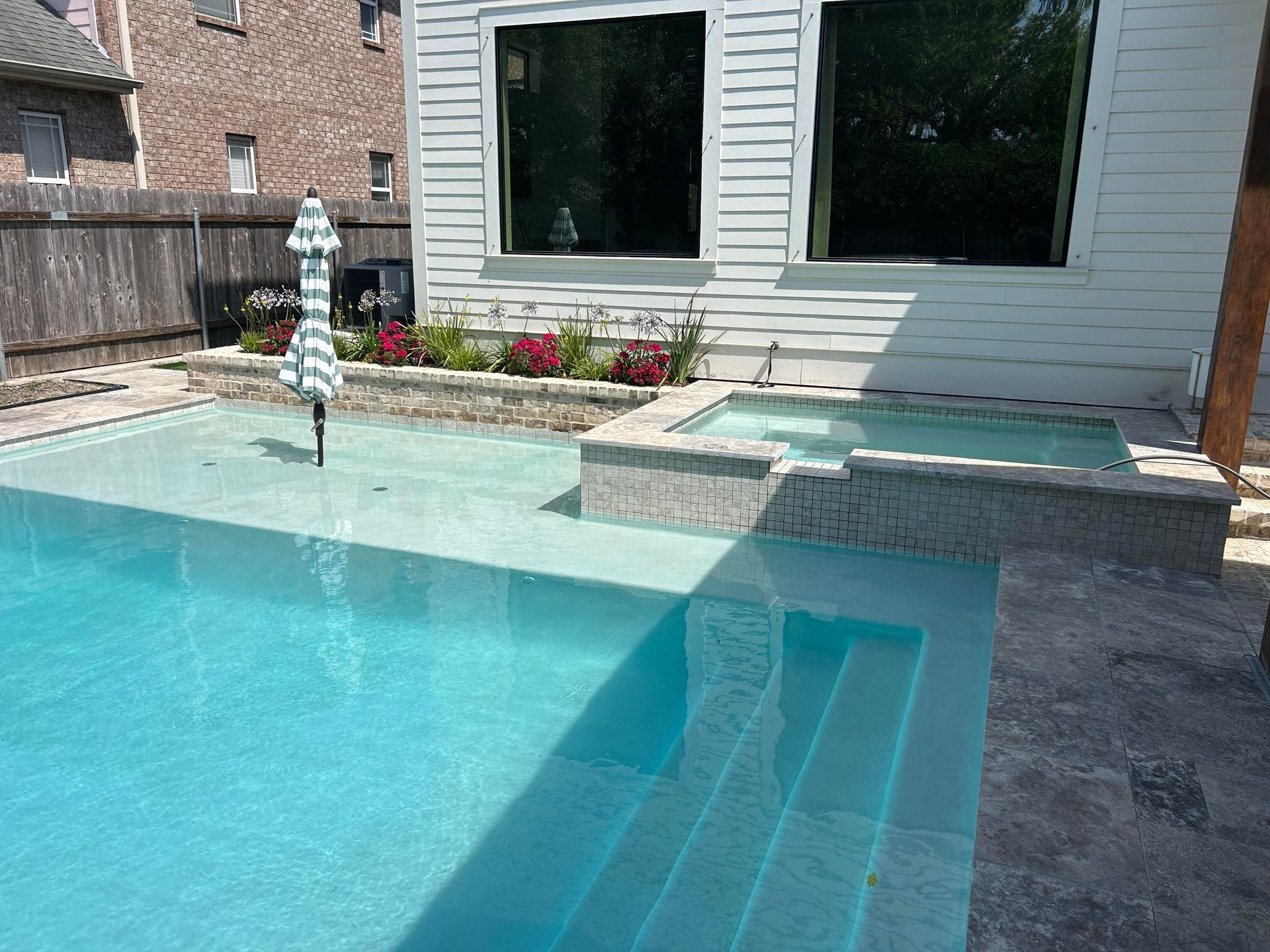 A turquoise backyard swimming pool with a stone hot tub and a small umbrella, set next to a white house with landscaping.
