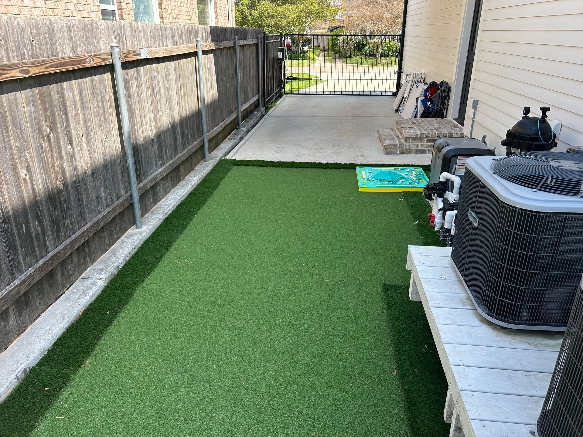 A side yard with artificial turf, a wooden fence, an HVAC unit on a white bench, and a concrete patio in the background.