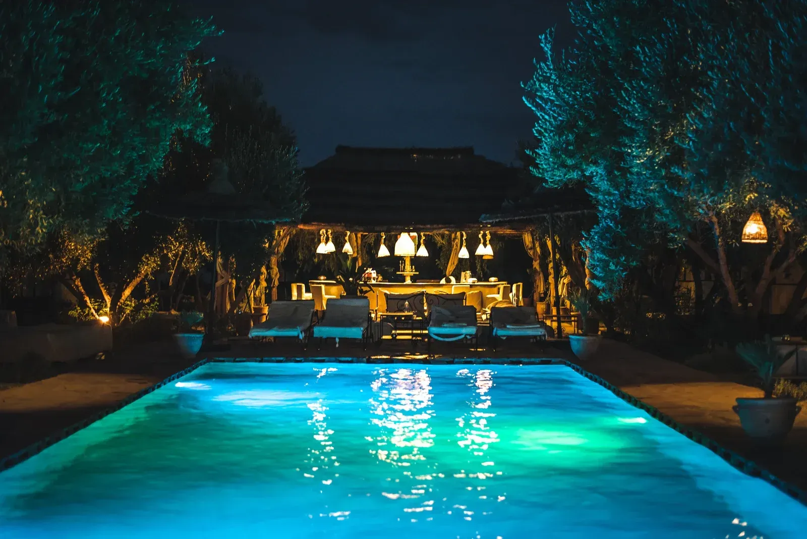 A large swimming pool is lit up at night with a gazebo in the background.