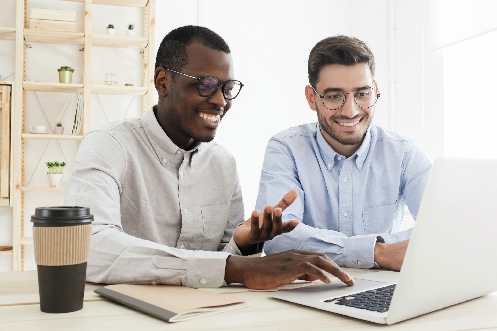 Two men wearing glasses looking at a laptop, smiling. One points at the screen. Coffee cup on desk.