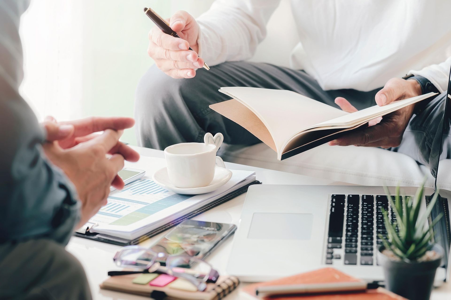Two people seated, reviewing documents at a table with laptop, coffee, and plant. One points with pen.