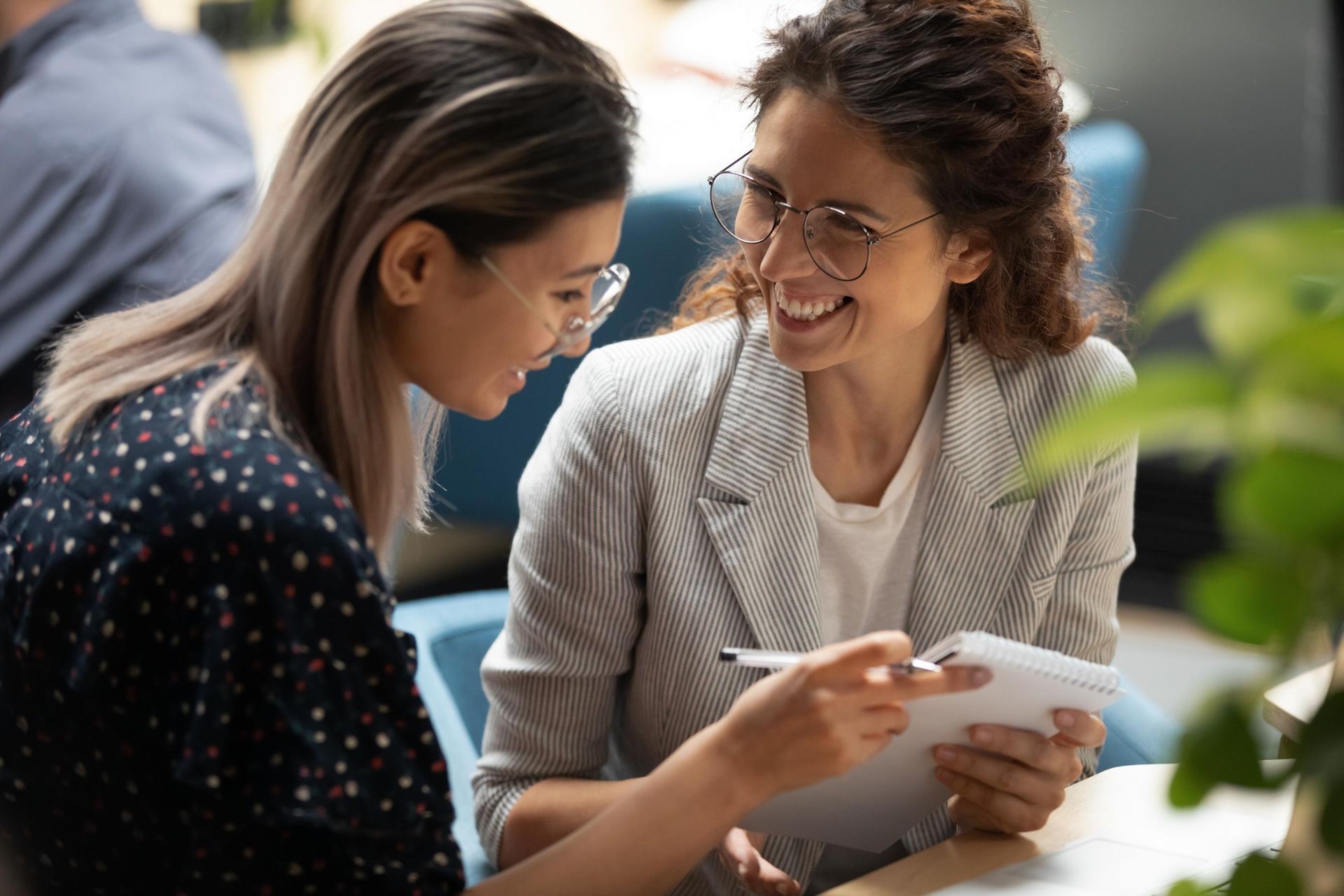 Two women smiling, looking at a notepad, indoors. One points while the other holds the pad and pen.