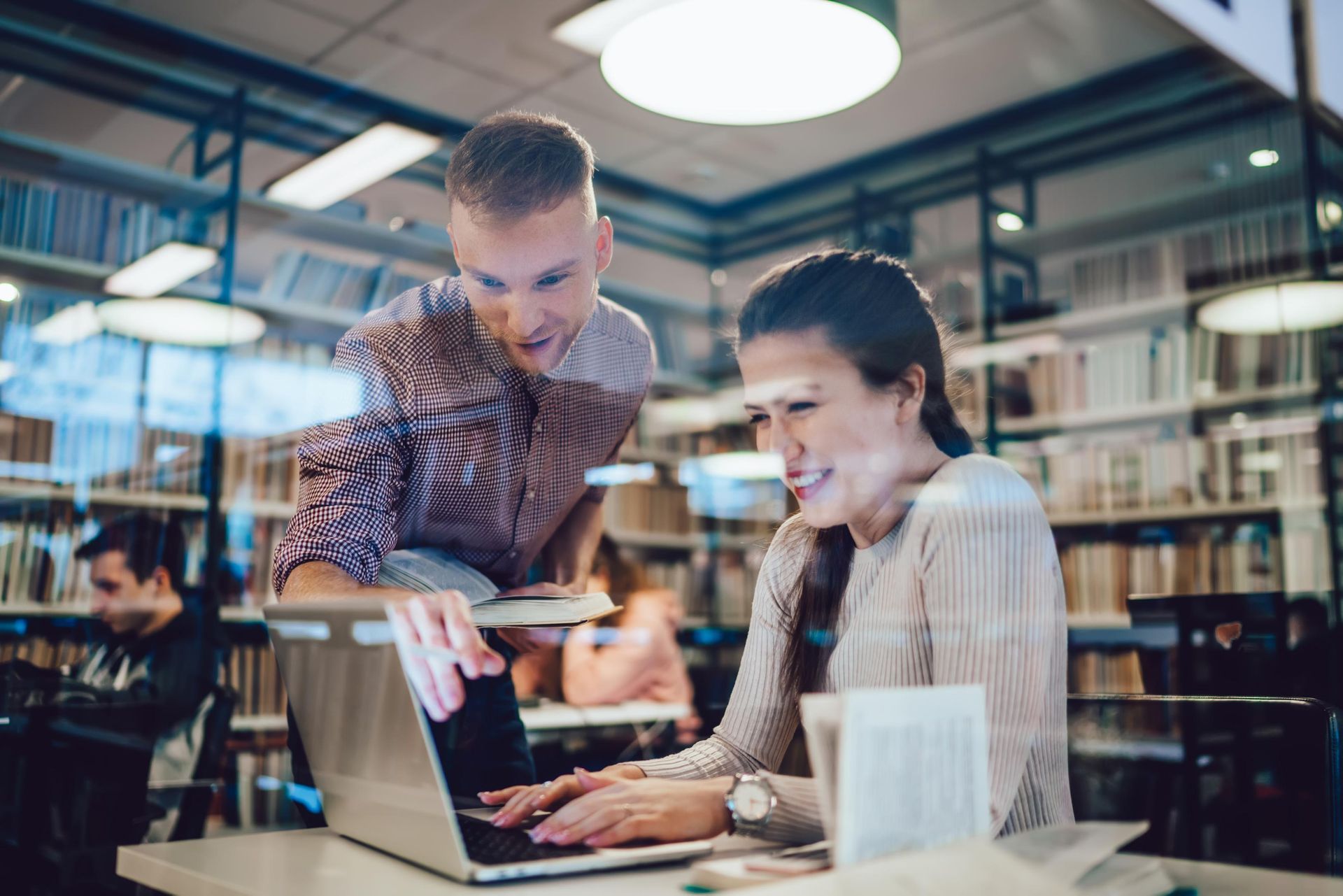 Woman using laptop; man leaning over, looking at screen, library setting.