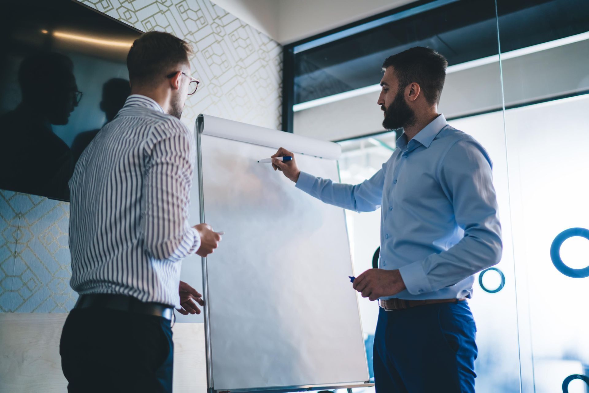 Two men in business attire at a whiteboard, one writing, the other observing in an office setting.