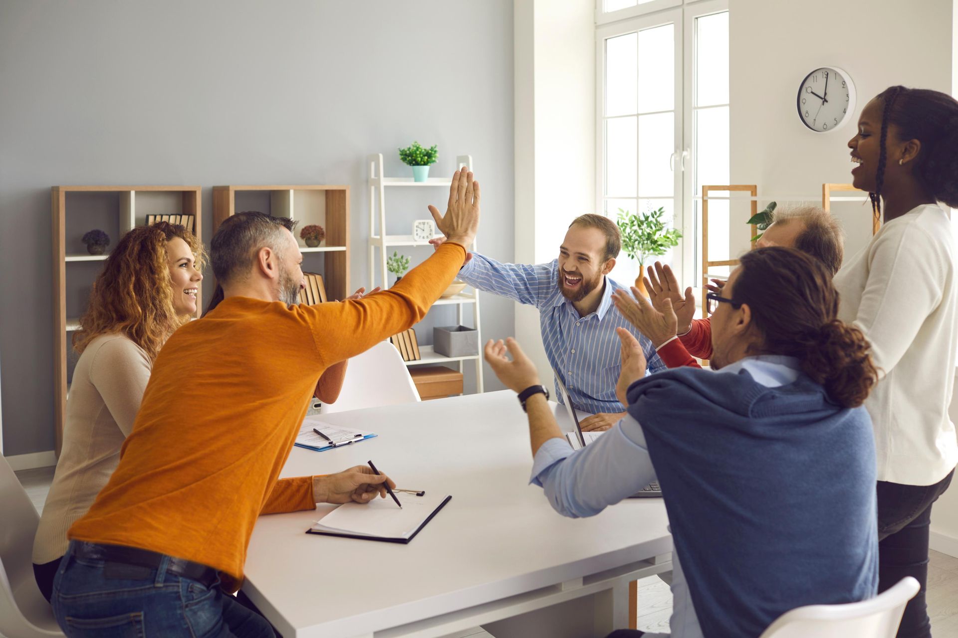 Team celebrates around a table, clapping and giving high-fives in a bright office setting.