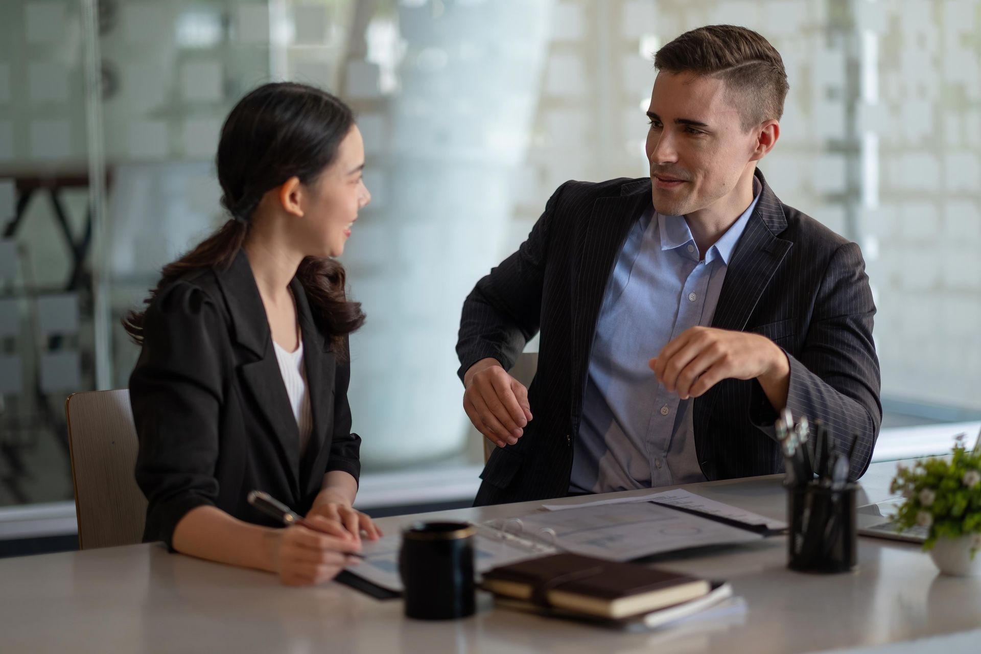 Woman and man in suits at a desk, conversing with documents, pens, and a small potted plant in an office setting.