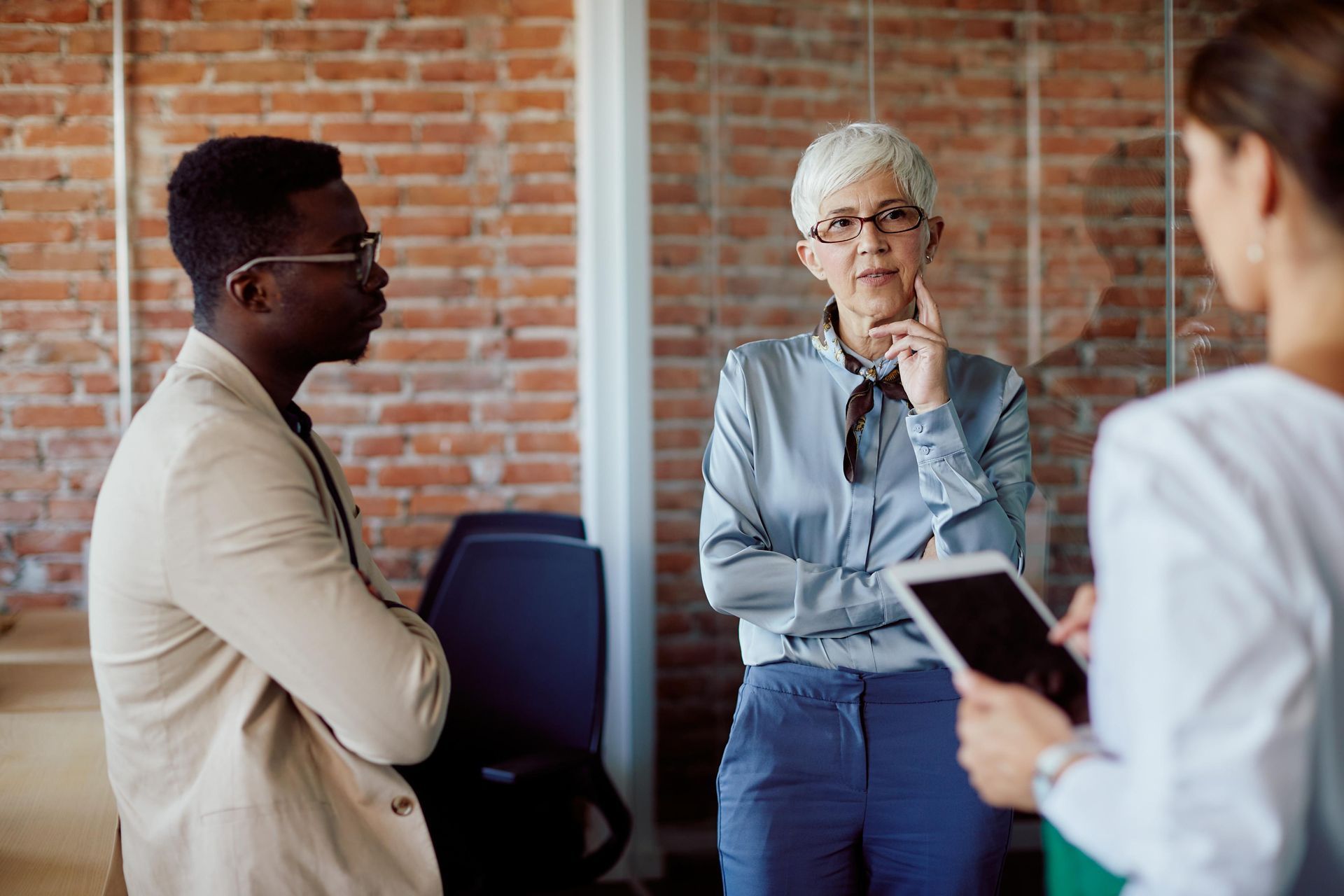 Three people in office setting, two men and a woman, discussing with a tablet. Brick wall backdrop.