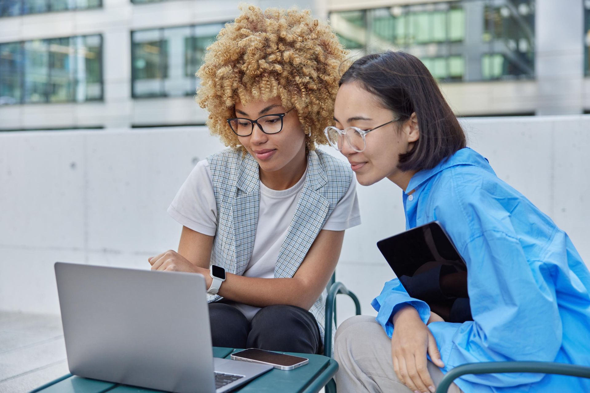Two people looking at a laptop outdoors. One with blonde afro and glasses points at the screen. The other holds a tablet.