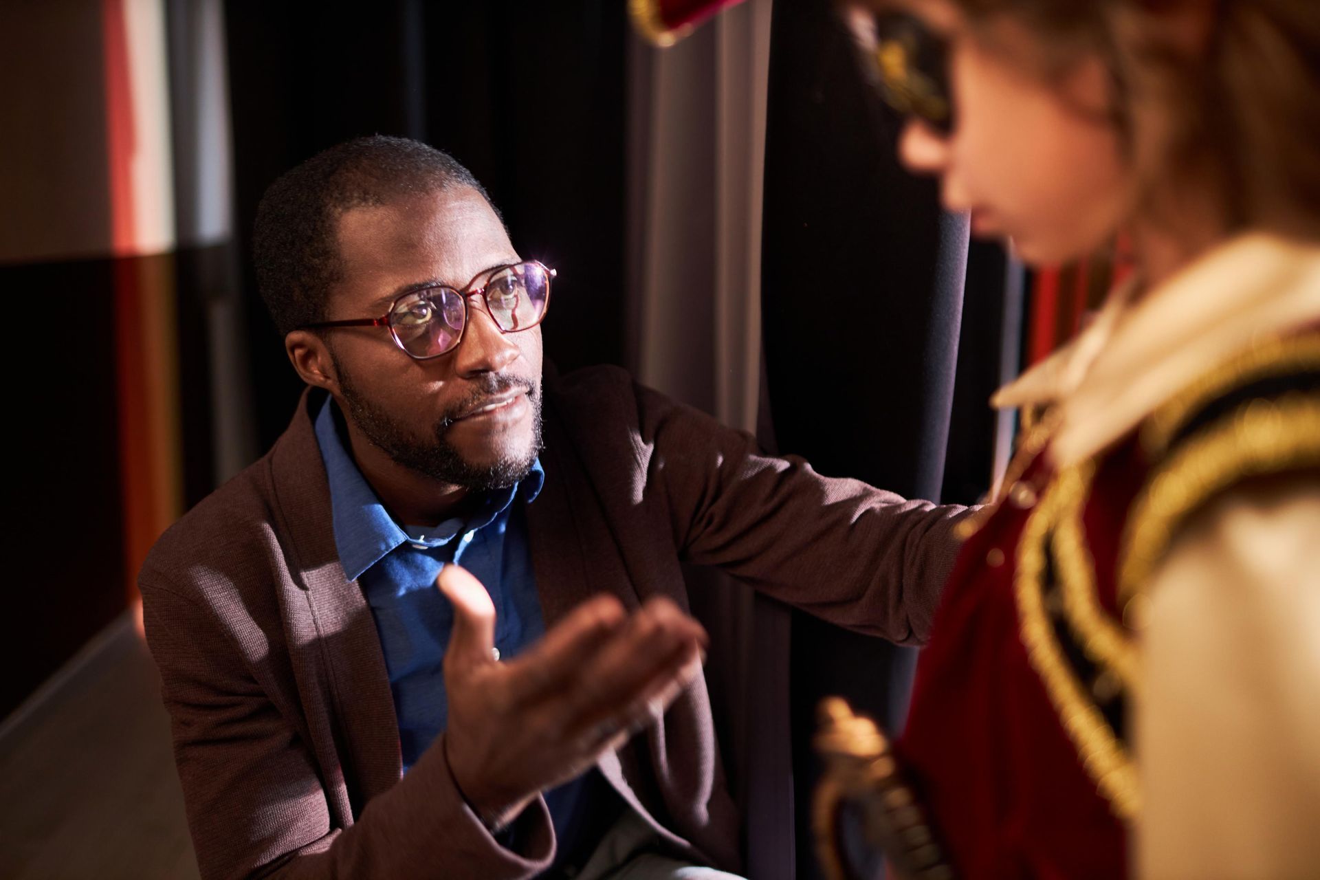 Man in glasses gestures, talking to someone in costume, indoor setting.