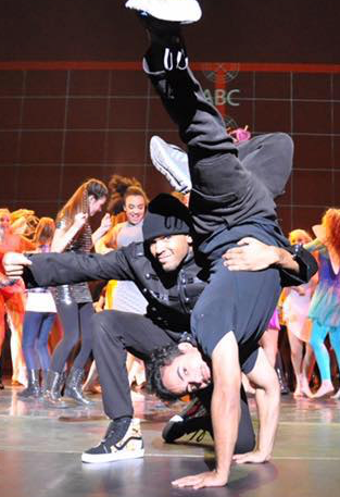 Two male dancers performing a handstand with one supporting the other's feet; stage with dancers in background.