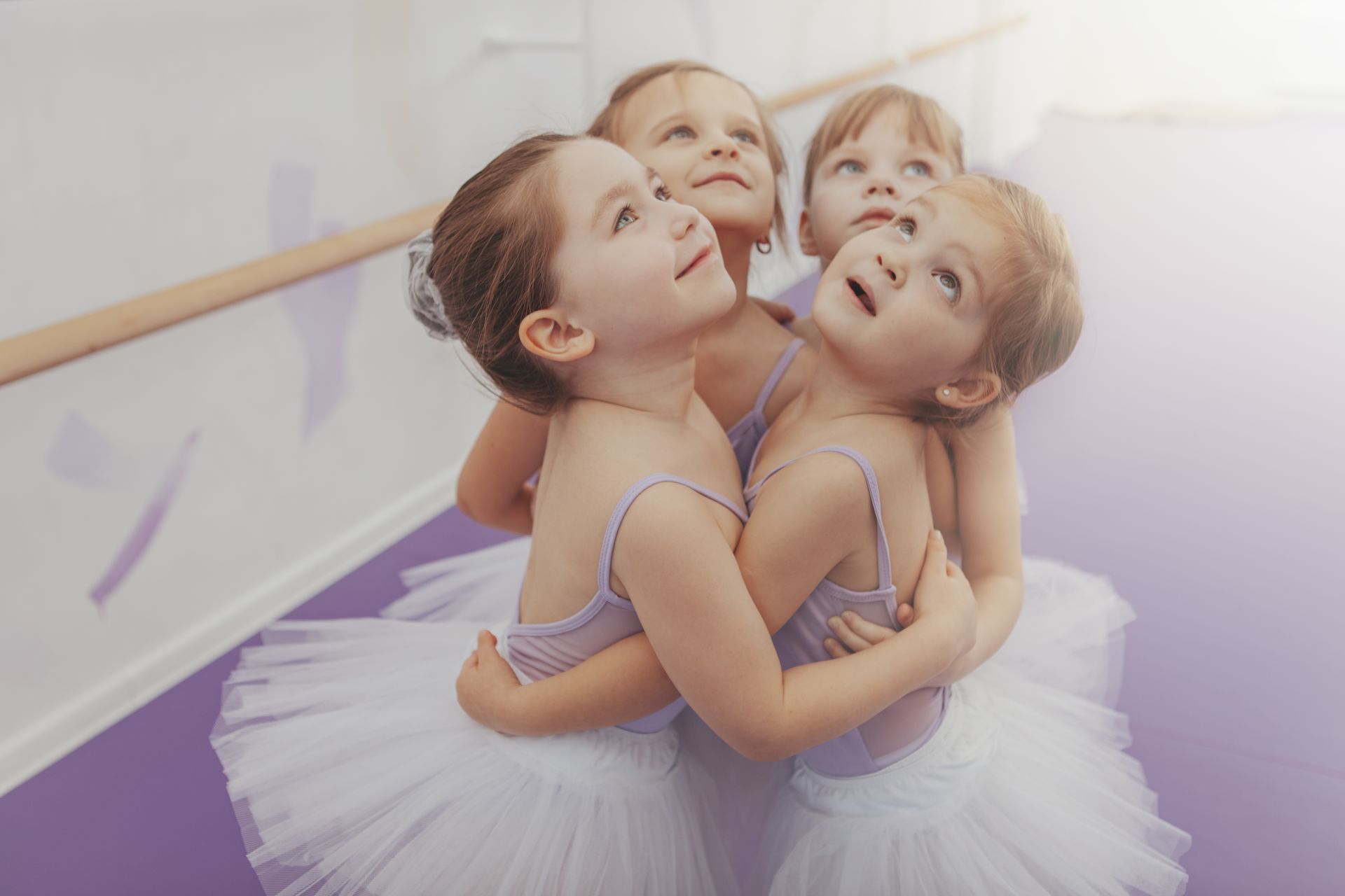 Four young ballerinas in white tutus and lavender leotards hug each other, looking upwards with surprised expressions in a dance studio.