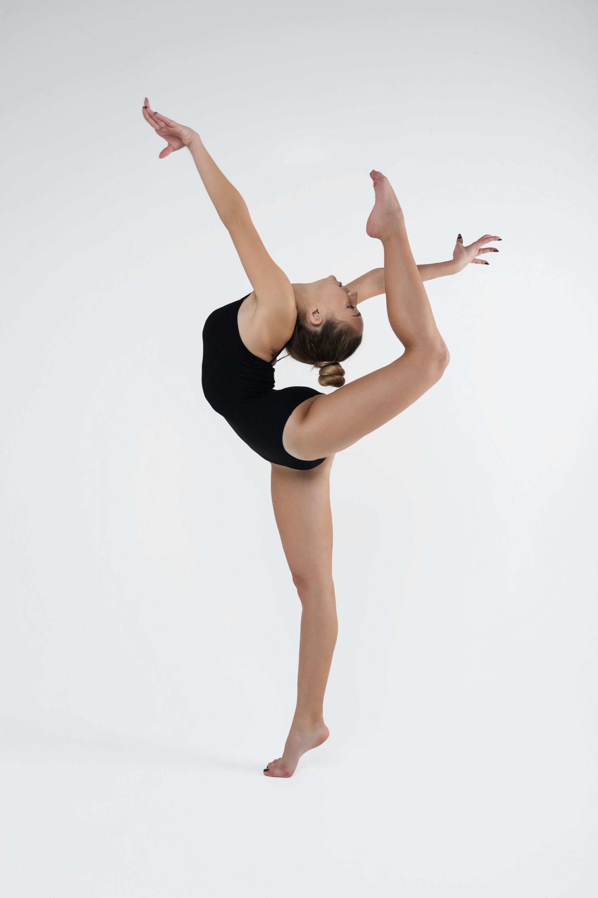A gymnast in a black leotard balances on one leg, arching her back and extending the other leg upwards, reaching for it with both hands.