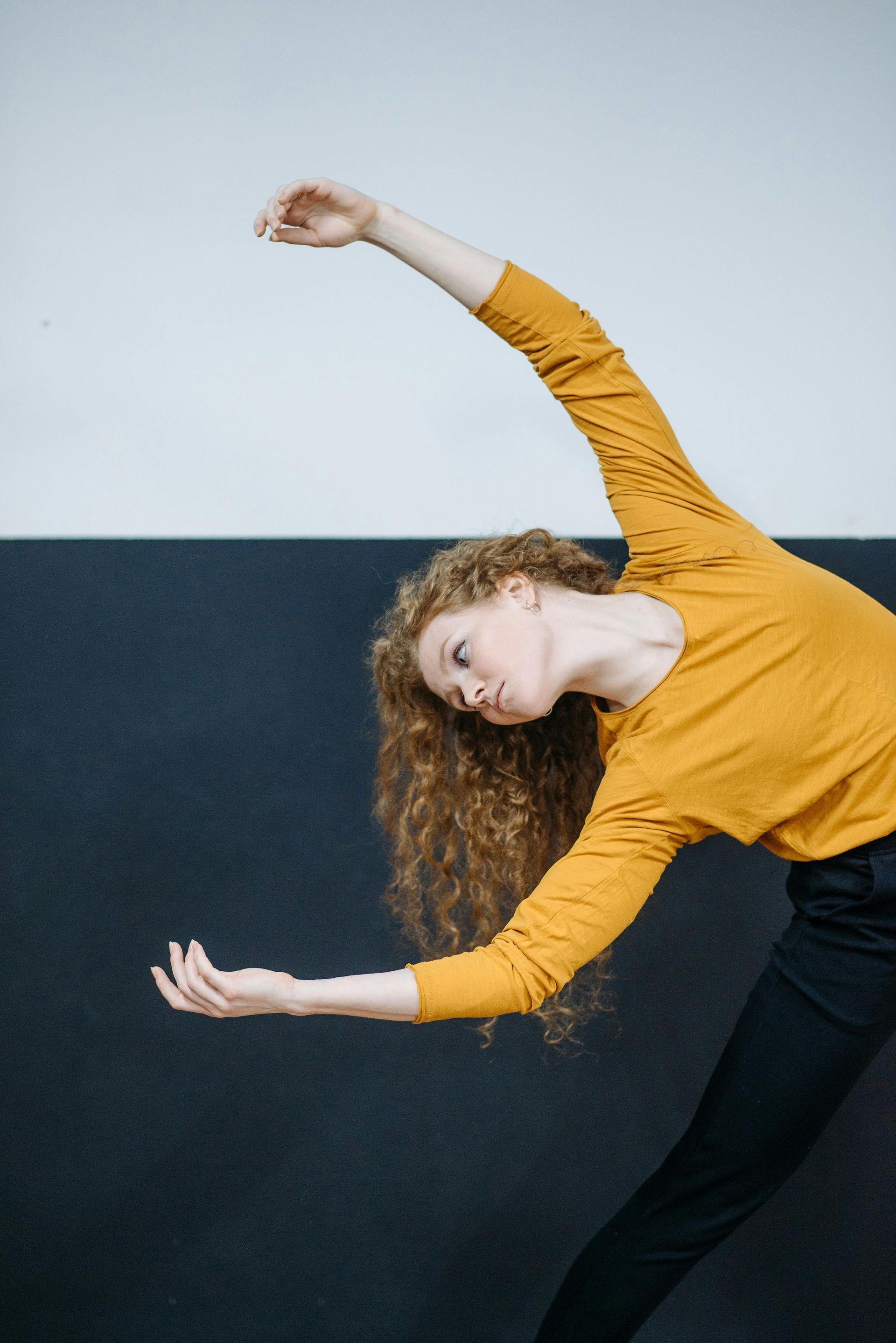 Woman with curly red hair wearing an orange shirt and black pants, bending to the side with arms outstretched against a two-tone wall.