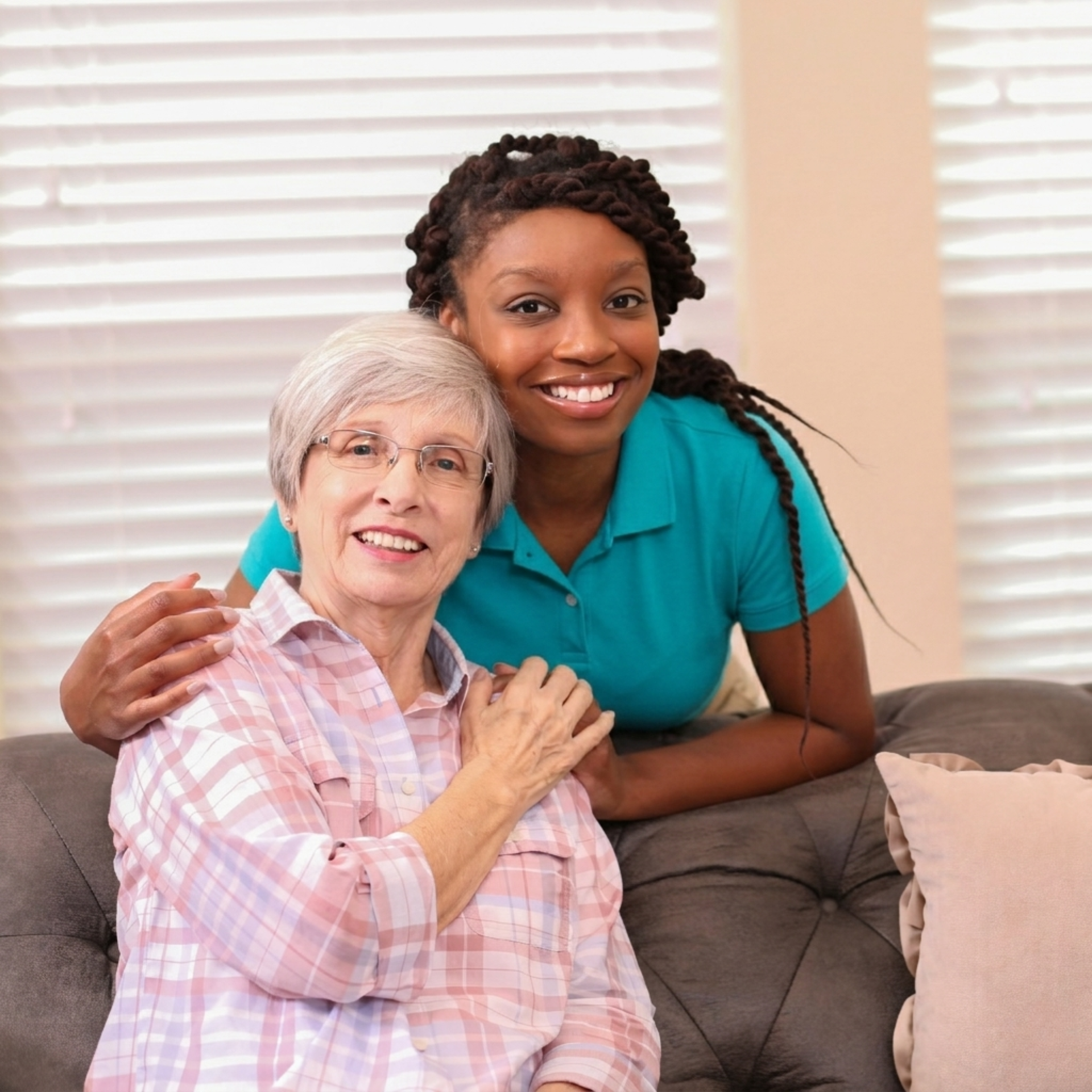 A caregiver with long braided hair in a teal polo smiles while standing behind a seated person in a plaid shirt.