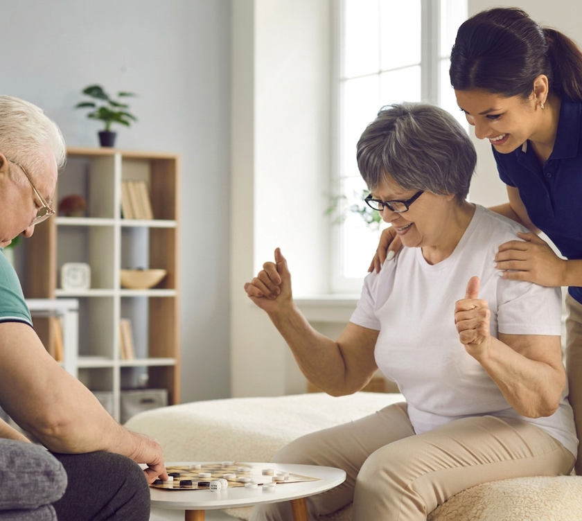 A caregiver stands behind a seated person, who is giving a thumbs-up while playing a board game with another person.