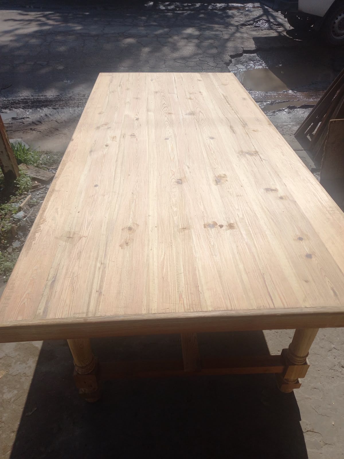 Mesa rectangular de madera para exteriores, expuesta al sol, con acabado claro y patas robustas.