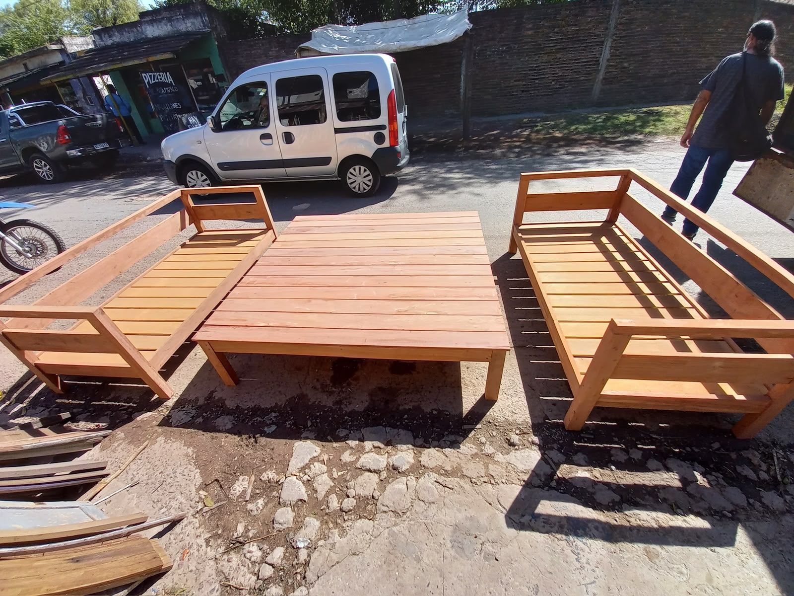 Plataforma de madera con asientos al aire libre en una calle pavimentada, con una furgoneta blanca y una persona al fondo.