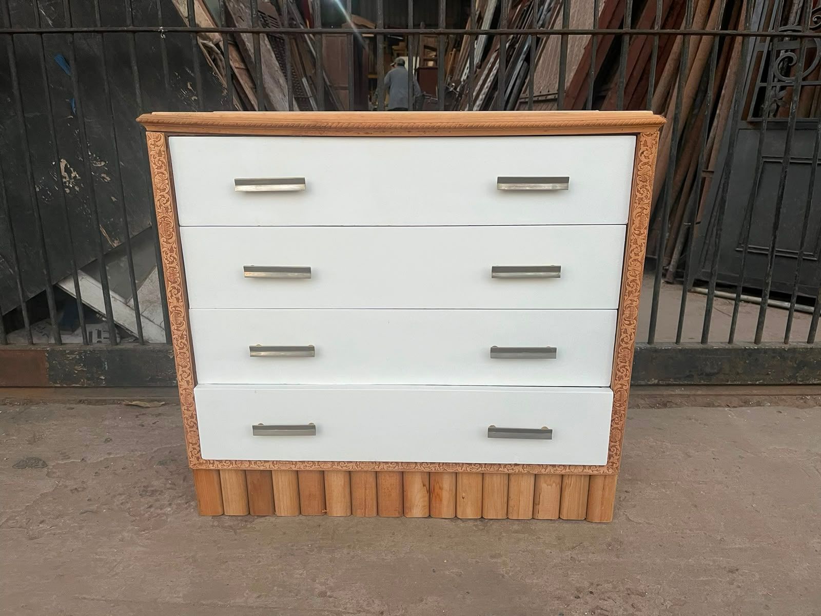 Cómoda blanca de cuatro cajones con detalles de madera, colocada al aire libre frente a una cerca de alambre.