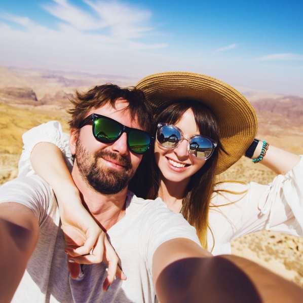 A man and a woman in sunglasses and hats smile during a sunny outdoor selfie in a desert landscape.