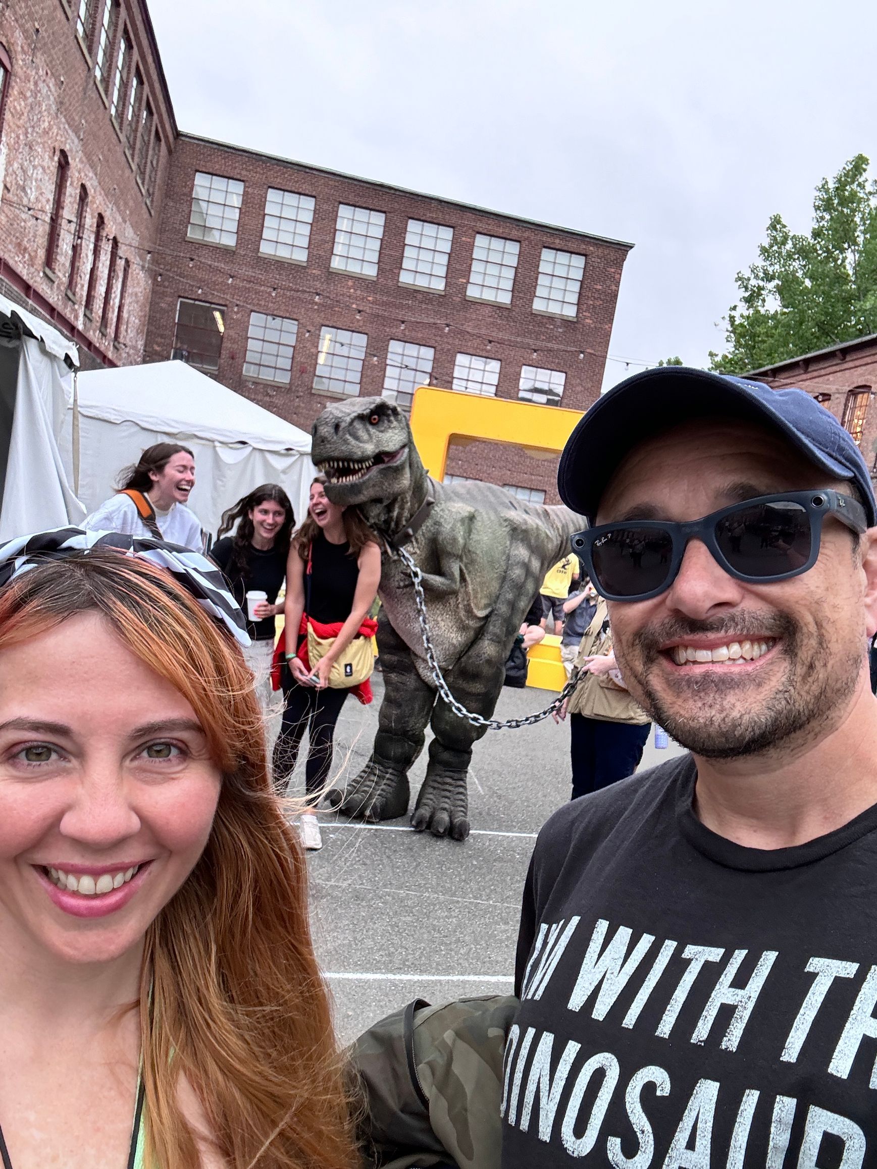A couple selfies with a dinosaur costume at an outdoor event; other people laugh in the background. Brick building in the backdrop.