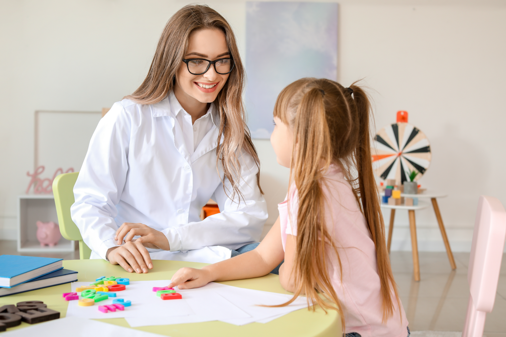 Une femme thérapeute portant des lunettes sourit à une jeune fille assise à une table avec des lettres colorées et un cahier d'exercices.
