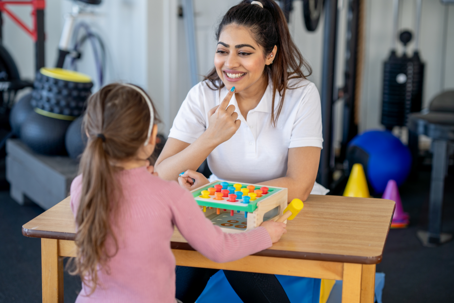 Une femme sourit à une fille qui joue avec un jouet de correspondance des couleurs à une table ; elles sont dans une salle de sport.