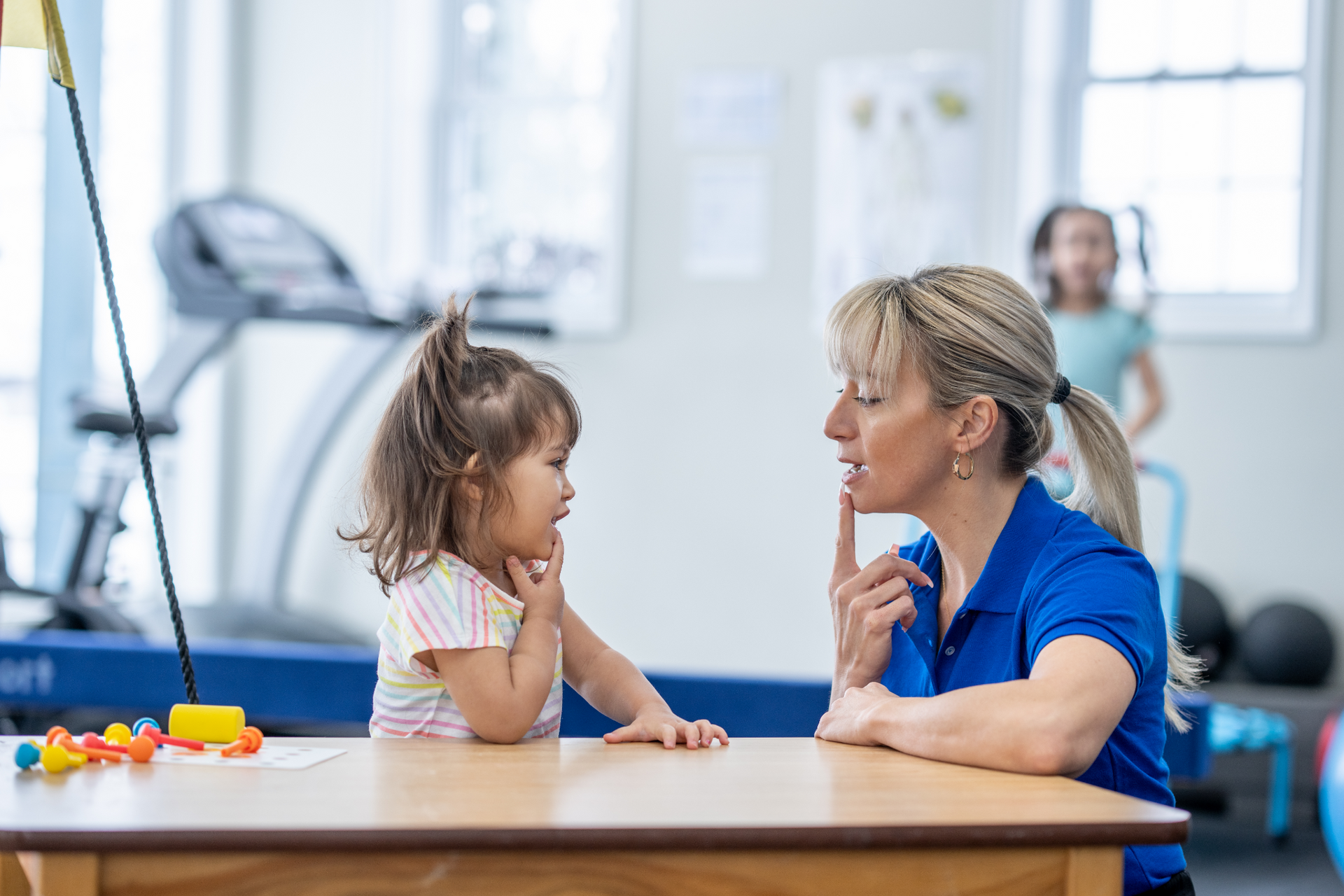 Une orthophoniste travaille avec un jeune enfant à une table. Une femme blonde fait un geste du doigt, tandis que l'enfant la regarde.