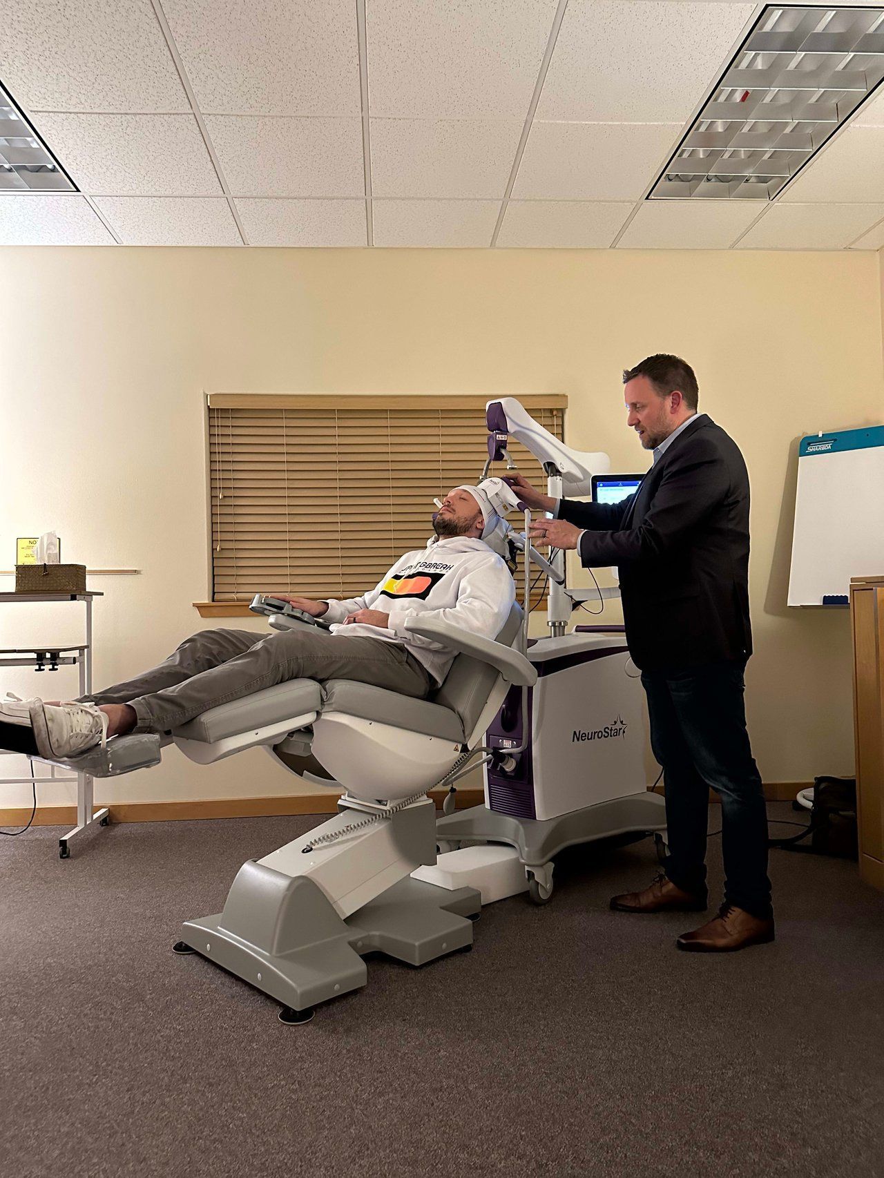 Man receiving treatment in a medical chair while another man adjusts equipment. The setting is a doctor's office.