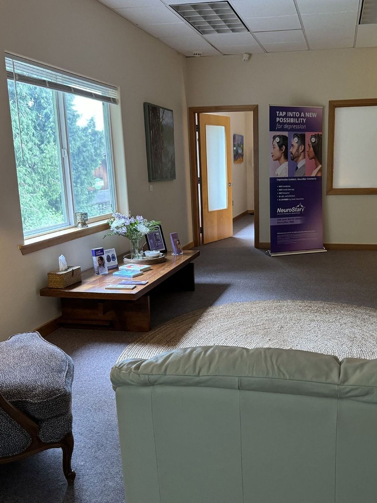 A waiting room with a low wooden table with flowers and brochures near a large window, couch, and a banner.