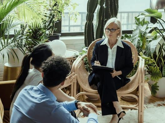 A therapist in a blazer sits in a wicker chair, taking notes while counseling a couple in a room with plants.