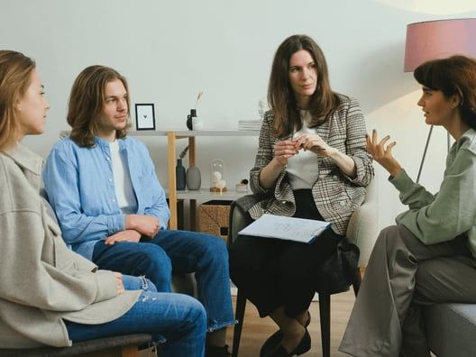 Four people in a circle; one therapist, three clients, in a counseling session. The therapist gestures while two clients listen.